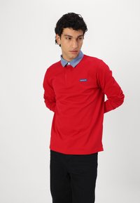 Red long-sleeve polo shirt with a chambray collar and blue logo patch. The fabric appears smooth, with a relaxed fit and cuffed sleeves.