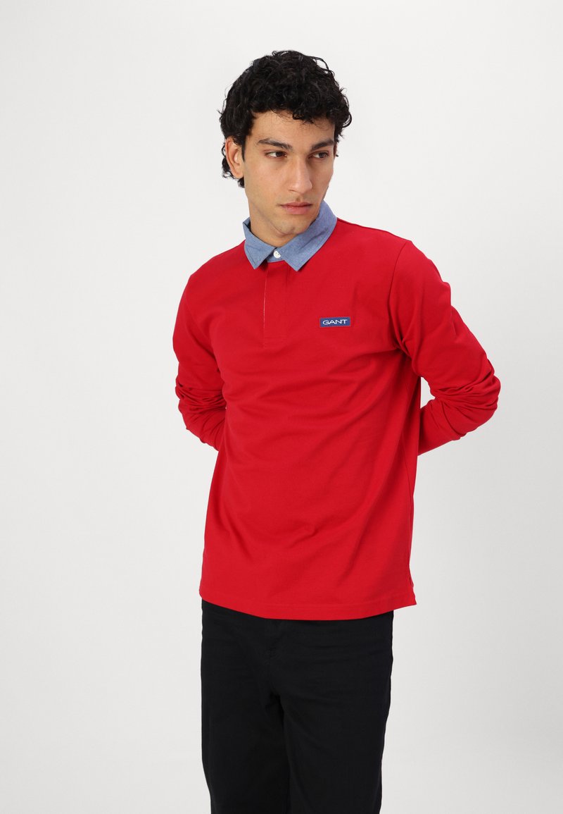 Rotes langärmliges Polohemd mit einem Chambray-Kragen und blauem Logo-Patch. Der Stoff wirkt glatt, mit einer lockeren Passform und umgeschlagenen Ärmeln.