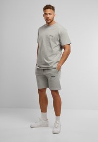 Grijze katoenen t-shirt en korte broek set met klein zwart logo. De korte broek heeft zijzakken en een trekkoord. Witte sneakers en crew-sokken.
