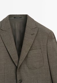 Brązowy żakiet z fakturowanej tkaniny, z klapami w stylu notch, z pojedynczym zapięciem na guzik i kieszenią na piersi. Wnętrze wyłożone gładką tkaniną.