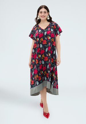 Kobieta uśmiechająca się, ubrana w granatową sukienkę w kwiaty z czerwonymi kwiatami i szarym wykończeniem, w połączeniu z czerwonymi szpiczastymi szpilkami i białymi kolczykami, stoi na białym tle.