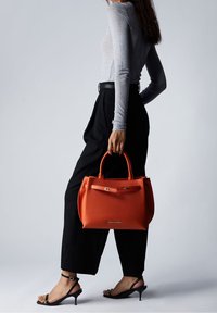 Sac en cuir orange au design structuré, avec des poignées doubles et un fermoir, complété par une finition texturée. Pantalons noirs à jambes larges et sandales à lanières.