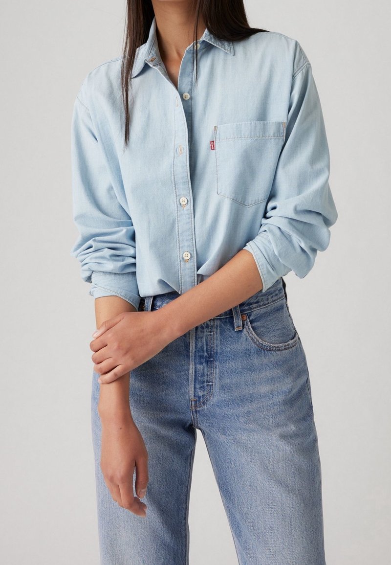 Femme portant une chemise en denim bleu clair à manches retroussées et un jean bleu taille haute, les mains croisées à la taille, devant un fond uni.