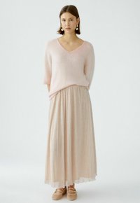 Lichtroze v-hals trui, zachte textuur; lange, geplooide lichtgouden rok met glans; beige platte schoenen met straps. Eenvoudig en elegant ontwerp.