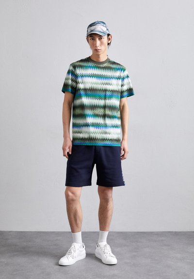 T-shirt κοντομάνικο με ζικ-ζακ σχέδιο σε πράσινες και μπλε αποχρώσεις, συνδυασμένο με ναυτικού χρώματος σορτς και λευκά sneakers με υφασμένες σόλες.