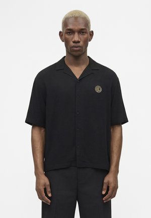 Mladý muž s krátkymi blond vlasmi nosí čiernu košeľu s krátkym rukávom zapínanú na gombíky s okrúhlym zlatým logom na hrudi, stojí pred bielym pozadím.