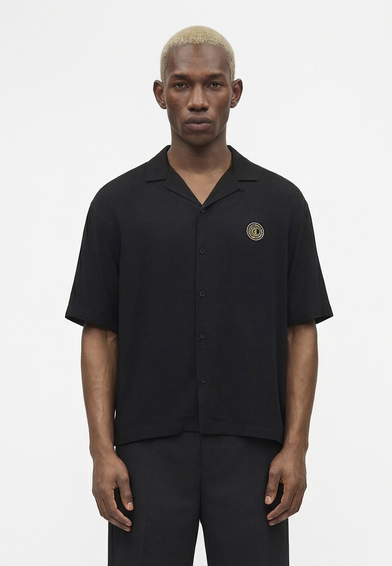 Νέος άνδρας με κοντά ξανθά μαλλιά φοράει μαύρο πουκάμισο με κοντά μανίκια και στρογγυλό χρυσό λογότυπο στο στήθος, στέκεται μπροστά σε λευκό φόντο.