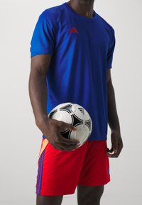 Blå sport t-shirt med röd Adidas-logotyp, kombinerad med röda shorts med orange detaljer och en svart Adidas fotboll i handen.