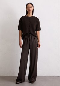 Lichtbruine gebreide korte mouw cardigan met knoopsluiting, gecombineerd met een losse bruine geplooide broek, wat een gestroomlijnde silhouet creëert.