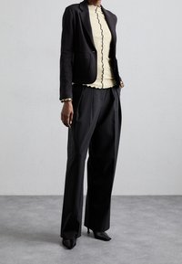 Černý blazer s klopami, kombinovaný se světle žlutým topem s volánkovým výstřihem a černým lemem, a volnými černými kalhotami.