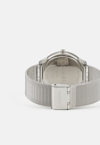 Reloj con correa de malla plateada y caja de acero inoxidable pulido. Presenta un diseño minimalista y un cierre marcado con "Calvin Klein."