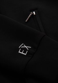 Sudadera negra con un bolsillo lateral con cremallera y el logotipo bordado "EX" en blanco en la manga. Textura suave, diseño minimalista.