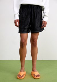 Black athletic shorts with elastic waistband, side pockets, contrast trim, paired with orange slip-on sandals. Smooth texture and casual design.