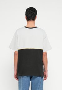 Κοντομάνικη μπλούζα με σχέδιο σε χρωματικά μπλοκ, με ανοιχτό γκρι στην κορυφή, μαύρο στο κάτω μέρος και κίτρινη διακριτική γραμμή κατά μήκος της ραφής.