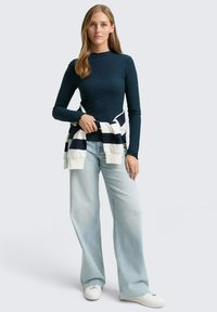 Marineblaues Ripp-Longsleeve mit einem gewellten Ausschnitt, kombiniert mit hellblauen, weiten Jeans und einem weiß-blauen gestreiften Pullover, der um die Taille gebunden ist.