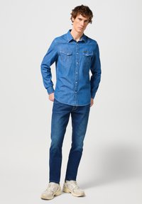 Camisa de denim en azul medio con mangas largas, botones de presión, dos bolsillos en el pecho y detalles de costura en contraste. Combinada con jeans azul oscuro.