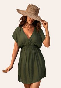 Vestido corto verde, ligero, con escote en V y mangas cortas, que presenta una cintura elástica y aberturas a los lados. Se lleva con un sombrero de paja.