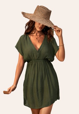 Vestido corto verde, ligero, con escote en V y mangas cortas, que presenta una cintura elástica y aberturas a los lados. Se lleva con un sombrero de paja.
