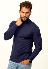 Navyblauer Strickrollkragenpullover mit gerippten Manschetten und Saum. Glatte Textur, figurbetontes Design. Getragen mit hellblauen Jeans und einer Uhr.
