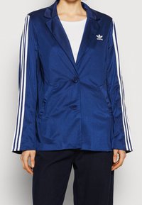 Sako z tmavě modrého saténu s jedním knoflíkem, dvěma předními kapsami a bílými třímístnými pruhy na rukávech. Na hrudi je umístěno logo.