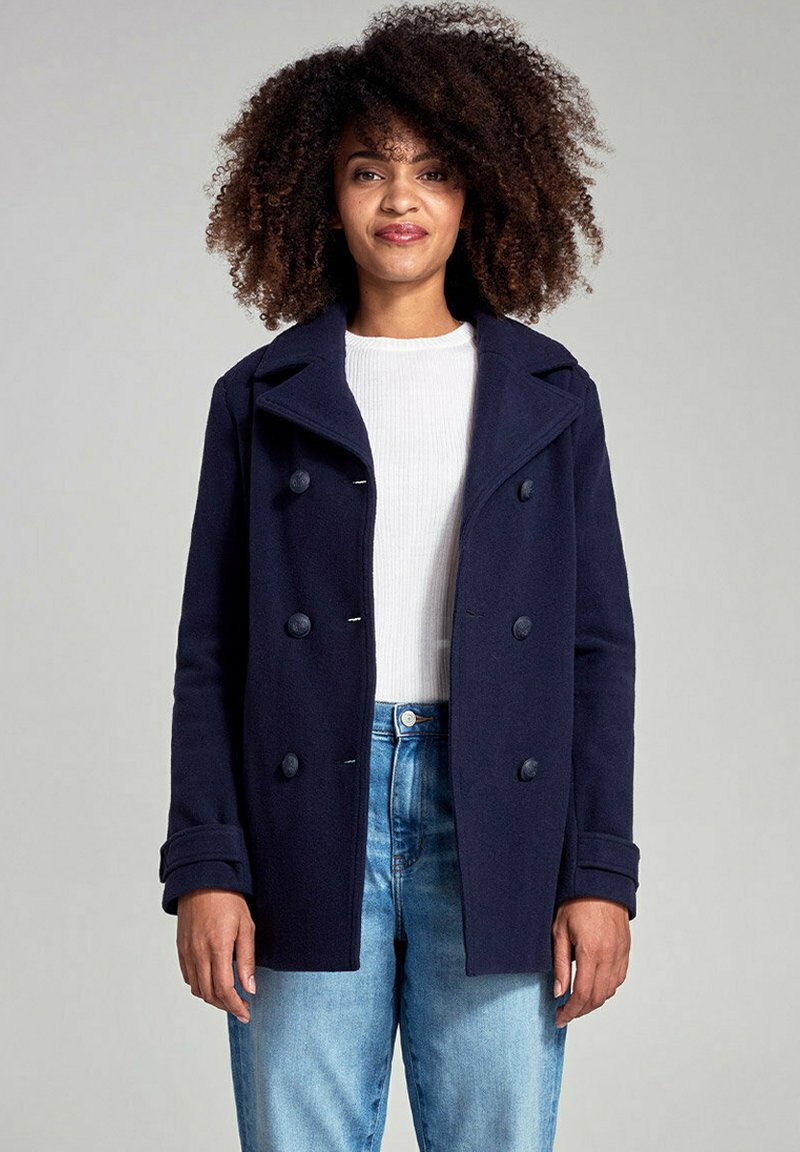 Pelliccia in lana blu navy con bottoni doppi, ampi rever e tasche laterali, abbinata a un maglione bianco a coste e jeans blu.