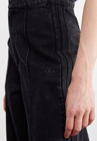 Črne denim hlače s tremi navpičnimi šivi na straneh, z vrezanim Adidasovim logotipom na levem stegnu.