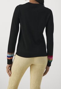 Kobieta ubrana w czarną bluzkę z długim rękawem z kolorowymi falbaniastymi mankietami oraz jasnoskalnymi, teksturowanymi spodniami, widziana od tyłu.