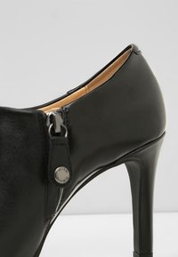 Chaussure à talon haut en cuir noir avec un bout pointu, détails de zip métalliques et texture lisse, dotée d'une doublure beige et d'un talon aiguille fin.