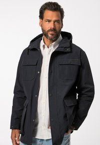 Tummansininen takki, jossa on huppu, useita etutaskuja, vetoketjusuljenta ja nappikiinnitys. Yllä vaalearaitaista paitaa.