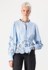 Blusa azzurro chiaro con bottoni, caratterizzata da un colletto arricciato, ricami floreali neri, vita elastica e maniche a sbuffo con polsini ondulati.