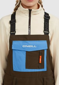 Bruine tuinbroeken met een blauwe zak en O'Neill merknaam, voorzien van verstelbare zwarte schouderriemen en een ritszak aan de zijkant. Fleece kraag zichtbaar.