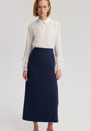 Sieviete valkā baltu blūzi ar garām piedurknēm, kas dekorētas ar kārtainām volānu aprocēm, un tumši zilu augsta vidukļa potītes garuma svārku, stāvot pret vienkāršu fonu.