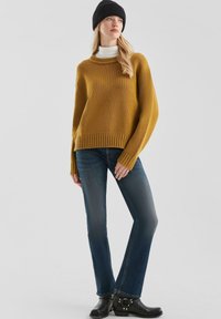 Przepiękny, oversize sweter w kolorze musztardowym z prążkowanymi mankietami i dołem, zestawiony z ciemnoniebieskimi dżinsami oraz czarnymi botkami za kostkę z ozdobnymi ćwiekami.