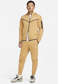 Tan Nike tracksuit with a zip-up hoodie and matching pants. Features black accents and logo. Model wears beige sneakers with black details.