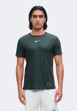 Homme aux cheveux noirs mi-longs, portant un maillot de sport Nike vert foncé avec des bords rouges et un short blanc sur un fond uni.
