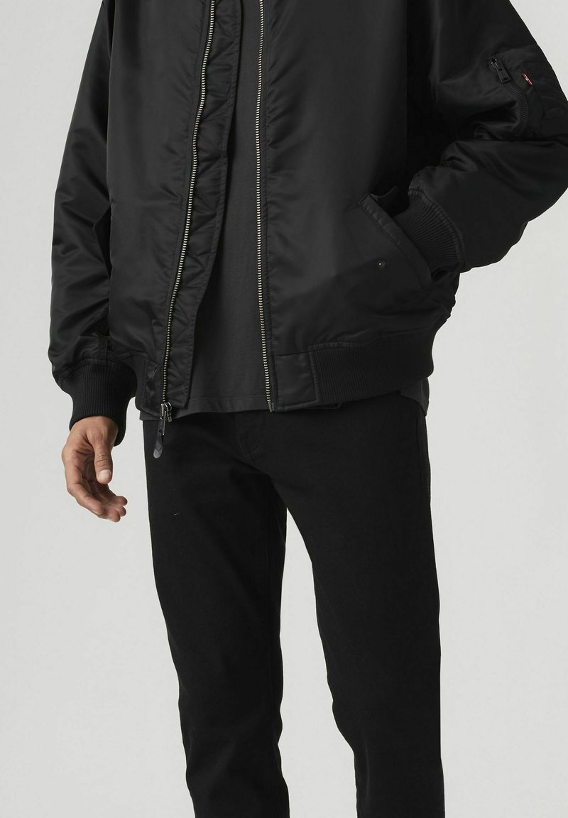 Homme portant une veste bomber noire avec poignets et ourlet côtelés, pantalon noir, et main dans la poche de la veste sur un fond uni.