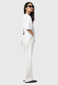 Witte oversized t-shirt met een open rug, gecombineerd met losse witte broeken en zwarte gestifte schoenen. Accessoires zijn onder andere een paar donkere zonnebrillen.