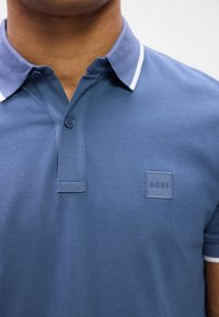 Polo shirt bleu en tissu texturé, doté d'un col classique avec des accents blancs et d'un patch logo carré sur la poitrine.