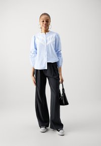Light blue striped button-up shirt with white lace accents, paired with dark wide-leg trousers and white sneakers. Holds a black handbag.