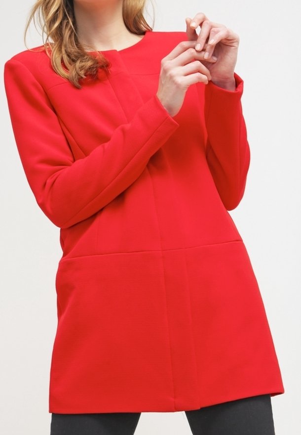 Rote Tunika mit langen Ärmeln, die eine strukturierte Form, glattes Material und Nahtdetails aufweist. Das Modell posiert mit gefalteten Händen vor sich.