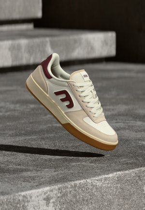 Crème en bordeaux sneaker met een gladleren bovenkant, geperforeerde neus, textuurkragen en gumrubberen zool. Veters en logo-details zichtbaar.