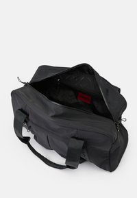 Bolsa de deporte negra con cremallera abierta que muestra un bolsillo interior con cremallera y una etiqueta roja con la marca "HUGO" en su interior.