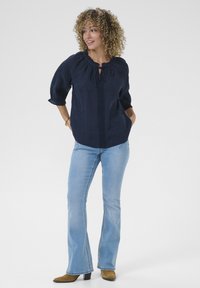 Navy blouse with a round neckline, tied at the front, featuring a textured panel down the center. Light blue flared jeans and brown ankle boots.