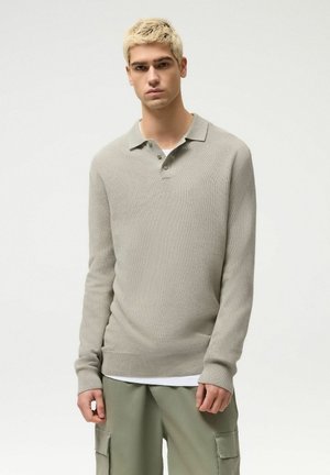 Młody mężczyzna o krótkich blond włosach, ubrany w beżowy sweter typu polo z dzianiny oraz oliwkowe spodnie typu cargo, na jednolitym białym tle.