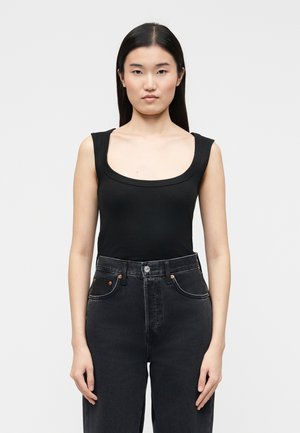 Egyenes fekete hajú nő fekete, ujjatlan, mélyen kivágott felsőt és magas derekú sötét farmert visel, és egyszínű háttér előtt áll.