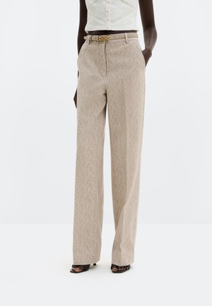 Személy bézs, magas derekú, széles szárú nadrágot visel finom halszálka mintával, övpánttal, fehér ujjatlan felsővel és fekete nyitott orrú magassarkúval.