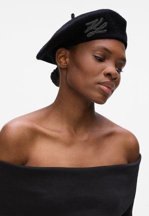Black wool beret featuring a textured silver accent. The subject has smooth skin and wears a black off-shoulder top.