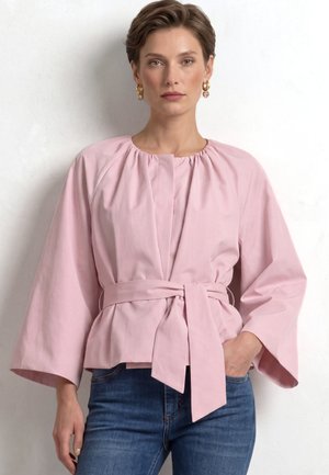 Mujer con blusa rosa claro ceñida con cinturón y mangas anchas y vaqueros azules, de pie frente a un fondo blanco.