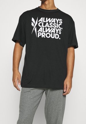 T-shirt noir avec un texte blanc en gras disant "TOUJOURS CLASSIQUE. TOUJOURS FIERS." Manches courtes et coupe décontractée, associé à un pantalon de survêtement gris.