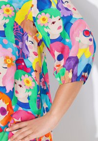 Tessuto colorato con un motivo di volti illustrati, caratterizzato da diversi colori di capelli e fiori. Il design include una manica a sbuffo e un polsino elastico.